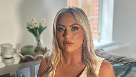 A woman with long blonde hair and blue eyes is looking directly into the camera, unsmiling. She is wearing a light-coloured sleeveless v-neck top. The background is out of focus but she is sitting on a grey solfa with light green cushions, behind her is a table with white flowers on it and a window. 
