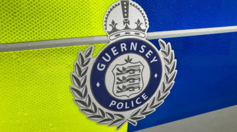 A Guernsey police logo with three lions and a wreath on a yellow and blue background.