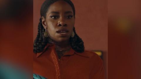 A woman with long dark hair and an dark orange top looks serious