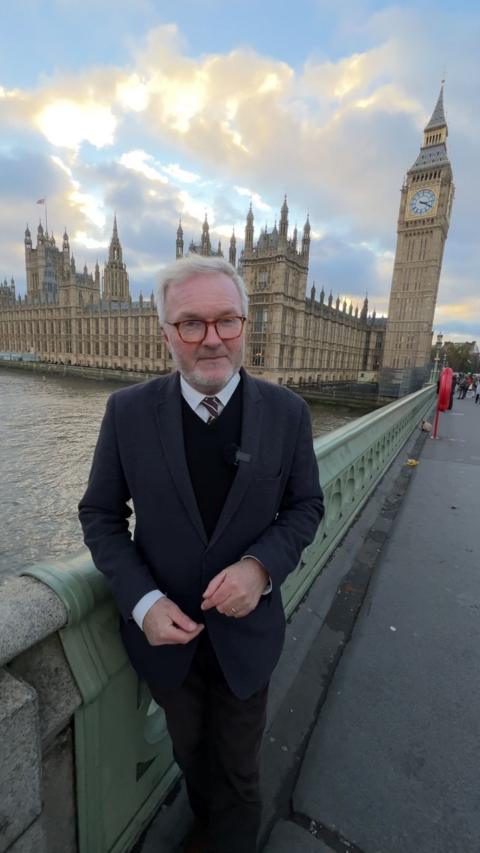 Political Editor Peter Henley wearing a dark suit and a shirt and tie standing on Westminster Bridge over the River Thames in London with the houses of Parliament and the Queen Elizabeth clock tower behind him. He is wearing square dark rimmed glasses.
