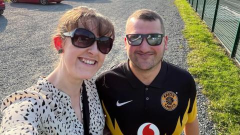 Menyw ifanc yn gwisgo gwisg wen a sbectol haul yn sefyll wrth ymyl dyn ifanc yn gwisgo crys pêl-droed yn tynnu hunlun wrth sefyll o flaen cae pêl-droed 3G.