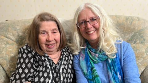 Mother and daughter, Maggie and Lucy are sitting, smiling on a sofa. Maggie is wearing a black and white top and Lucy is wearing a pale blue jumper with a blue, pink and green scarf.