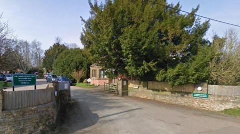 The entrance to Carrdus school - there is a wooden gate on one side of a driveway. There is also a green sign with a the school's name on it.