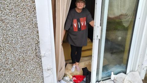 Claire Wheeler, wearing a t-shirt, leggings, a cap and odd socks, stands at the open glass sliding door of her house. There are shoes and clothes piled up by the doorway and a pile of sandbags just outside the door.