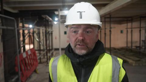 A man wearing a hard hat and hi-vis jacket stands in a building site