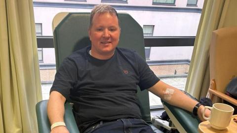 Ryan is sitting in a green leather hospital chair. He is wearing a navy t-shirt and jeans and is smiling at the camera. He has a hospital bracelet on his right hand and a canula in his left arm. Behind him is a window which has light green curtains and a white hospital building can be seen outside. 