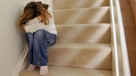 A child with mid-length brown hair sitting on a cream carpeted staircase with her arms wrapped around her head, which is resting on her knees. She is wearing a white jumper, blue jeans and pale pink socks.