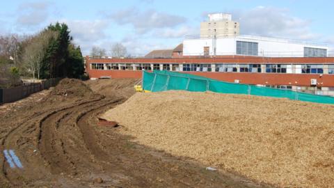Enabling works being carried out on the extension to Kettering General Hospital's maternity unit, showing mud on the ground, green barriers, a building behind it and stone on the ground. 