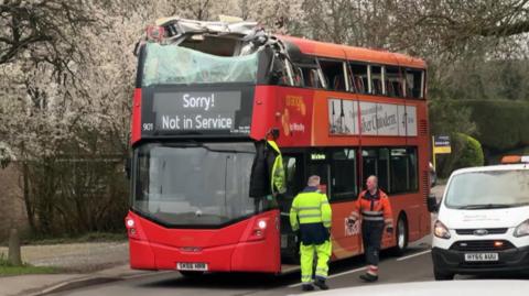 A red and orange double-decker bus is pictured stopped in the road. The upper deck is significantly damaged following a crash with a bridge. A message reading "Sorry! Not in Service" is being shown on the bus