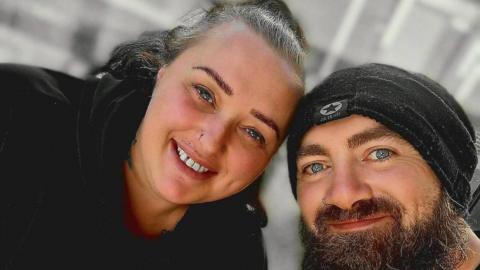 Llinos Hughes and her partner Paul in a selfie. Llinos has blue eyes and smiles at the camera, she is wearing a black hoodie. Paul also has blue eyes and a dark beard and wears a black hat.
