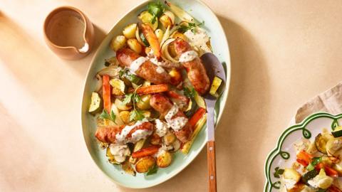 A blue plate full of sausages with roasted potatoes, carrots, parsnips, onion, courgette, and herbs.