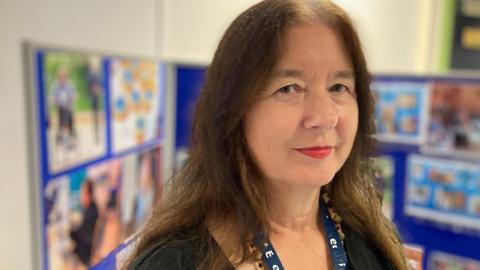 Helen Fairweather looks slightly over her shoulder toward the camera. She is smiling. She has long brown hair and wears red lipstick and a black cardigan with  a black and white vest underneath.