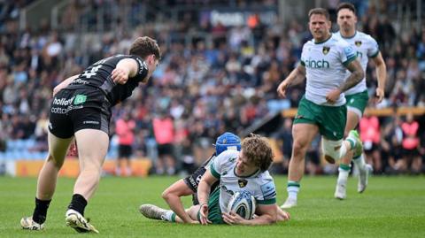 A rugby player in white celebrates having scored despite being tackled by an opponent with a blue scrum-cap. Three other rugby players look on with a stand blurred in the background.