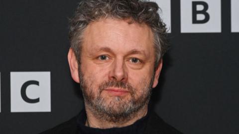 Michael Sheen, dark hair and beard, staring straight-faced at the camera