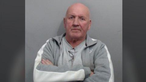 A man, standing in front of a grey background. He is bald and has his arms crossed. He is wearing a grey t-shirt and grey zip up jumper.
