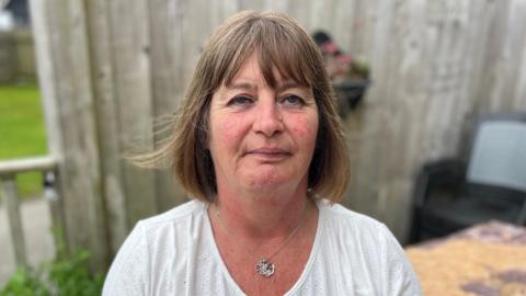 A woman in her 50s is sat on a garden bench with a fence behind her. She has mid length brown hair in a bob hair cut, and is wearing a white top with a silver necklace. She has a passive expression. 
