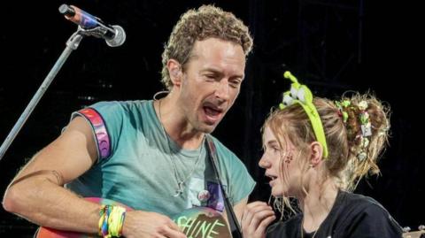 Chris Martin is standing close to Isla and is singing to her as he plays guitar. She is looking pleased and shocked. She has her long blonde hair piled in a bun which is decorated with flowers and beads and she is wearing a fluorescent head band. Martin is wearing a green T-shirt and has lots of festival bands wrapped around his wrist.