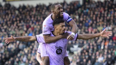 Lincoln City's Hackett celebrates a goal with his arms stretched wide. Captain, Darikwa, has jumped on his back in celebration and smiles as he looks forward. They are both wearing Lincoln's lilac away kit. The background is blurred but full of fans.