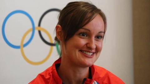 Susan Egelstaff smiling in front of the Olympic rings logo. She is wearing a bright red athletic jacket with a black zipper and a hint of a white and blue visible near the collar.