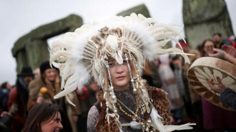 Revellers attend the winter solstice celebrations at Stonehenge, a person is in centre of the frame wearing a feathered headdress.