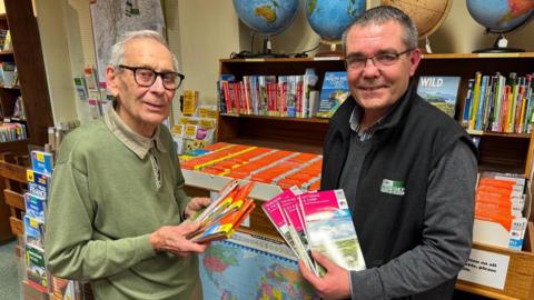 Two men are standing together and smiling at the camera. They are both holding a stack of maps and are standing in front of a bookshelf filled with maps