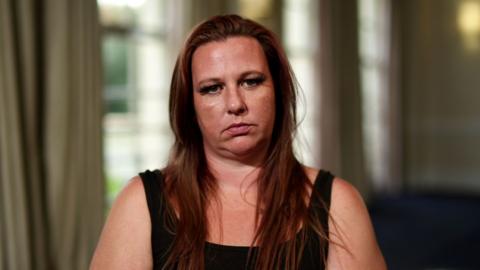 A woman wearing a black tank top and long reddish brown hair sits and stares at the camera.