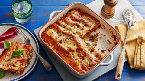 A square oven dish on a white board with a serving spoon, filled with butter bean and kale cannelloni, with a portion on a plate to the side. 