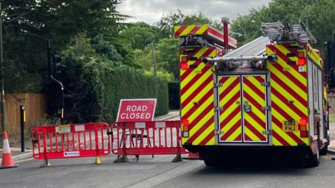 A fire appliance parked in College Road, Epsom, beside a road closed sign and two red plastic barriers blocking the road.