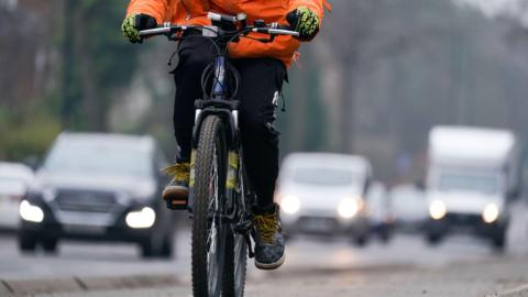 A person is on a bicycle on a road, with out of focus traffic behind. We can only see the person from waist down but they're wearing an orange waterproof coat and black and lime green cycling gloves. The bike wheels have a fairly chunky tread.