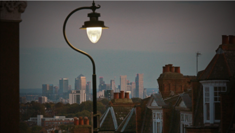 A general view of London at night, looking down a street of terraced houses. High-rise buildings can be seen in the distance and there is an old-fashioned street lamp in the foreground