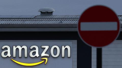 A photo from 2023, which is a close-up of an Amazon logo sign on an Amazon warehouse in Coventry. A red stop sign is visible in front of the factory.