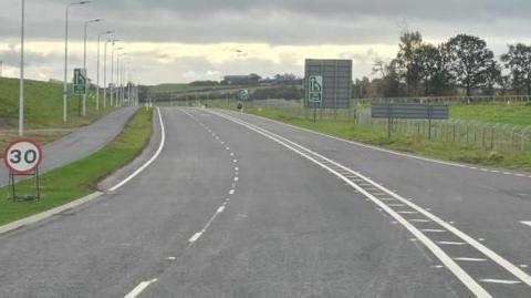 A section of road between Newby West Roundabout and the new Cummersdale Roundabout on the B5299 at Dalston Road in Carlisle. The road and its markings are new with a temporary 30mph speed limit sign to the side. On the left is a grass embankment, on the right a flat area of grass and a few trees.