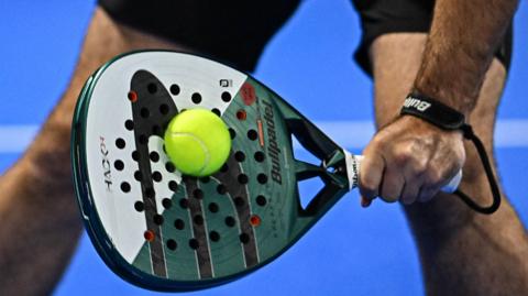 We can see a padel racquet being held by a man near his knees. It's connecting with a padel ball as he returns a shot. The racquet is a solid piece of equipment, as opposed to having strings like a tennis racquet would.