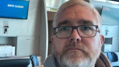 Jon Shipman. a man with white hair and a full beard, stands looking forlorn in an airport with a screen behind him bearing the words 'gate closed'. He is wearing black glasses.