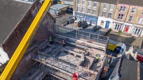 A large yellow crane lifts a steel beam into place at the top of a bombed-out cinema in Hull. Workers in hi-vis vests can be seen guiding the beam in place. The site is surrounded by scaffolding.