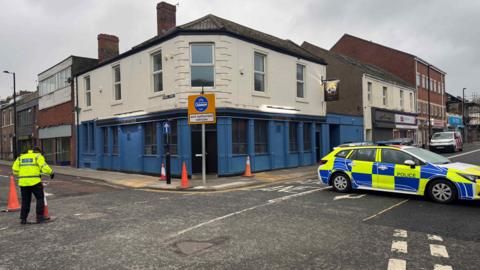 A blue and white public house and a row of shops. A police car is parked blocking the road and a police officer in hi-vis is stood on the road.