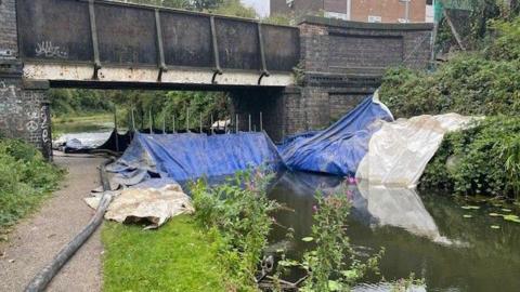 Walsall Canal, with pipes and tarpaulin across it to assist clean up after a toxic spill, underneath a bridge at the top of the frame