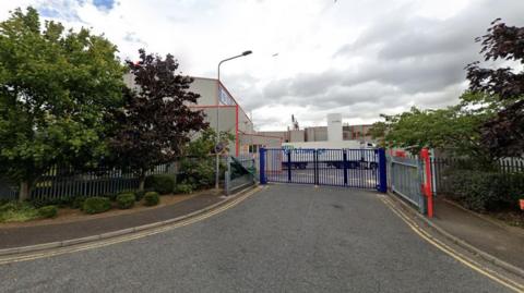 A view of a factory in an industrial estate taken from the road. Blue gates are across the entrance and a HGV is in the background.