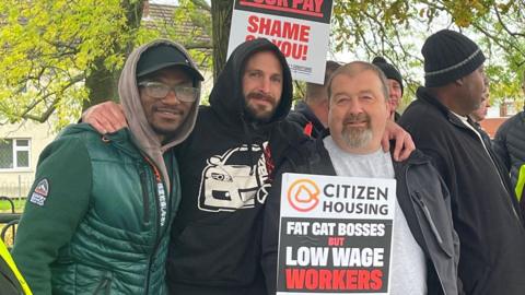 Three men holding placards stating "Fat cat bosses but low wage workers". ONe is wearing a green coat, the second is in a black hoodie and the third is in a grey t-shirt.