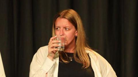 Ruby Tapp wearing a black top with a white shirt over the top of it. She is drinking from a glass which is in her left hand. She has shoulder-length brown hair and is looking to the left.