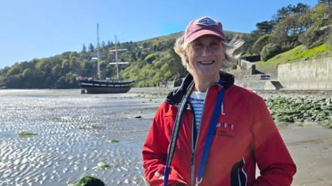 Penny Bielich has fair short hair and is wearing a faded red cap with a diamond logo on it. She is smiling and wearing a red coat with a blue dog lead hanging around her neck. She is standing on the beach on a sunny day, with the tall ship beached in the background on the left and a tree-lined hilly coastline beyond. 