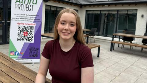 A teenage girl with long blonde hair, sitting at an outdoor bench and smiling at the camera. She is wearing a maroon T-Shirt.