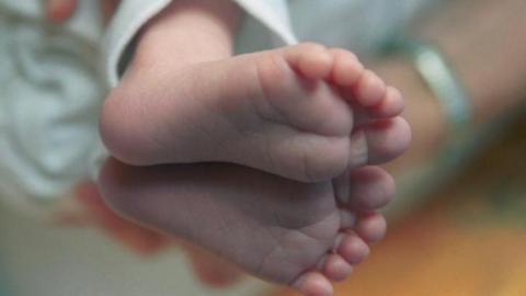 A newborn babies feet. In the blurred background, an adults arm with a silver bracelet on it can be seen cradling the baby.
