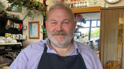 A man with short grey hair and a matching beard is standing in a cafe beside a large, black, Gaggia coffee machine. He is wearing a black apron over a lilac patterned shirt.