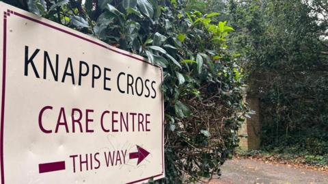 A sign which says Knappe Cross Care Centre This Way against a green hedge