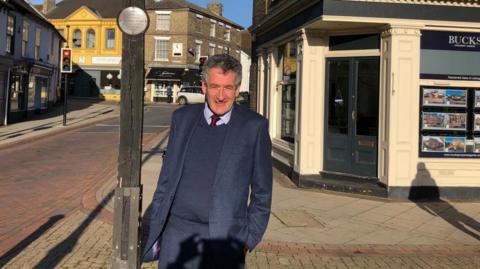 Peter Prinsley is standing in the Market Place in Stowmarket in front of an estate agents on the corner of a street. He is wearing a blue suit and jumper with a red tie. 
