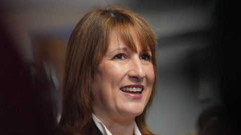 Chancellor Rachel Reeves smiles in a headshot. She wears a high-necked white shirt with a black jacket. Her light brown hair is work loose with a long fringe.