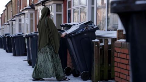 A woman moves a black bin, she wears a long green dress, it is a snowy residential street with lots of black bins outside red semi-detached houses.