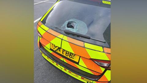 An orange and yellow marked PSNI car. The photo is taken from behind the car and the window has been damaged, with a hole in it.  