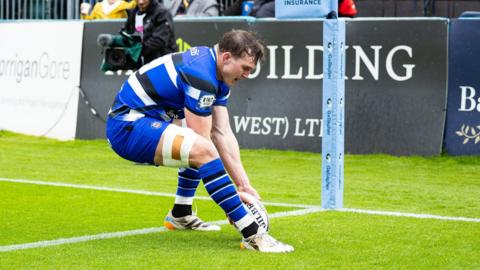 Ted Hill, wearing a blue shirt with black and white stripes, placing the ball over the try line to score near the corner flag during a game between Bath and Newcastle.